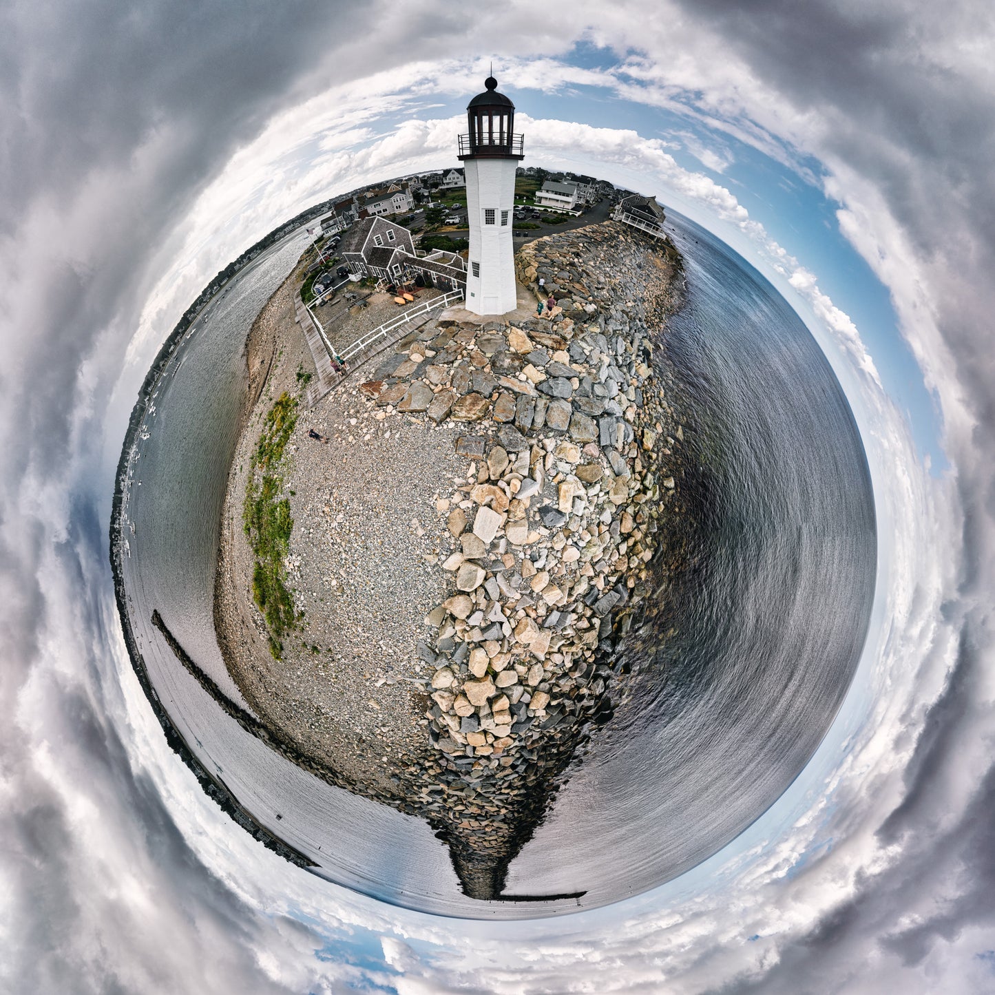 360-degree “tiny planet” image of Scituate Light on a rocky breakwater, forming a small circular world of stone, ocean, village houses, and dramatic swirling clouds.