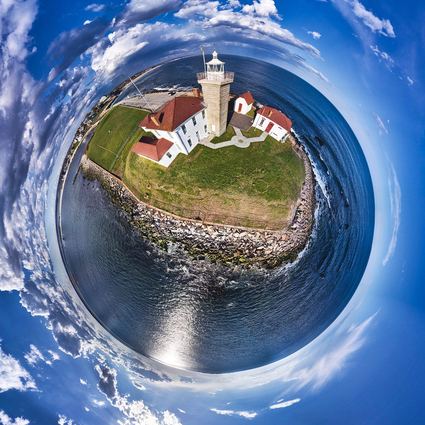 A 360-degree panoramic photograph of Watch Hill Lighthouse in Rhode Island, featuring white buildings with red roofs, a stone seawall, and dramatic cloud formations over deep blue waters.
