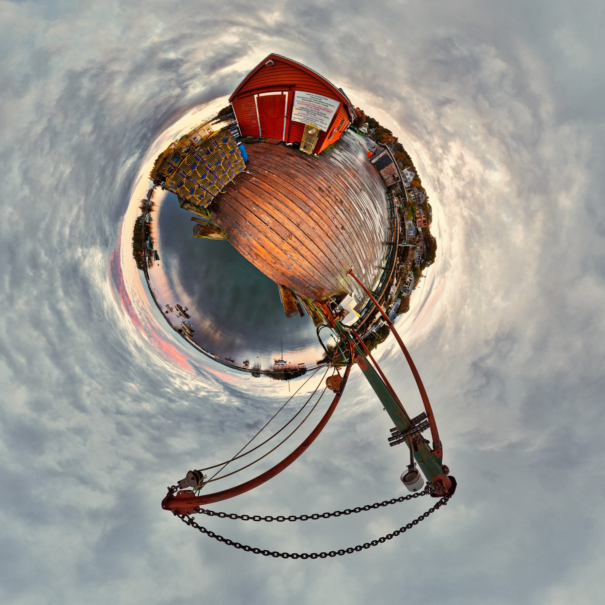 360-degree “tiny planet” image of the Five Islands, Maine working waterfront, showing a red lobstermen’s shed, stacked lobster traps, wooden dock, and hoist over a circular harbor under swirling gray clouds.