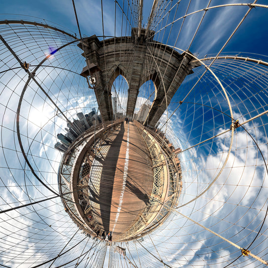 A 360-degree panoramic photograph of the Brooklyn Bridge, featuring radiating suspension cables, wooden walkway, gothic archways, and Manhattan skyline curved against a bright blue sky with clouds.