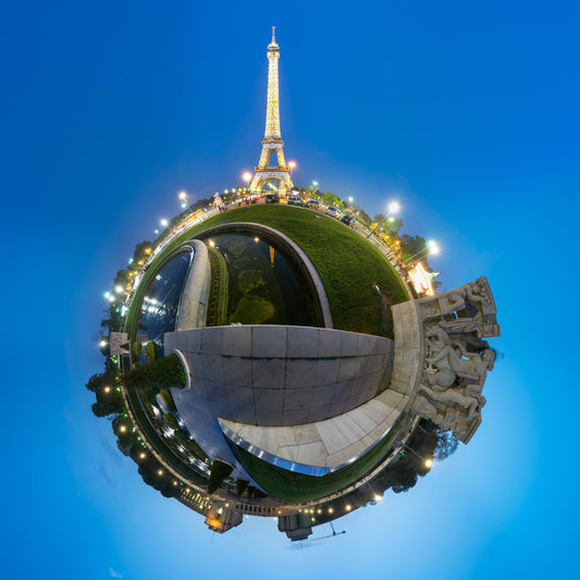 Spherical panoramic photograph of the illuminated Eiffel Tower at twilight, rising above the Trocadéro gardens and fountains, creating a perfect circular world against a deep blue sky.