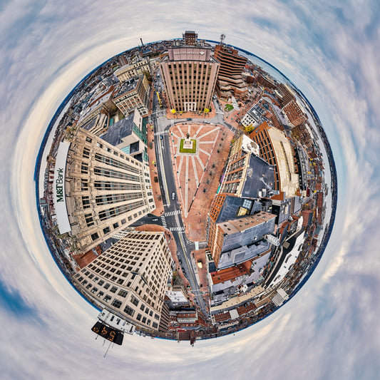 360-degree “tiny planet” image of Monument Square in Portland, Maine, with brick plaza and monument at the center, surrounded by leaning downtown buildings, streets, and a circular soft blue-gray sky.