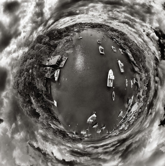 Aerial black and white 360-degree panoramic photograph of New Harbor, Maine, depicting boats anchored in calm waters, surrounded by a rugged shoreline, all encased within a sphere under a dramatic sk