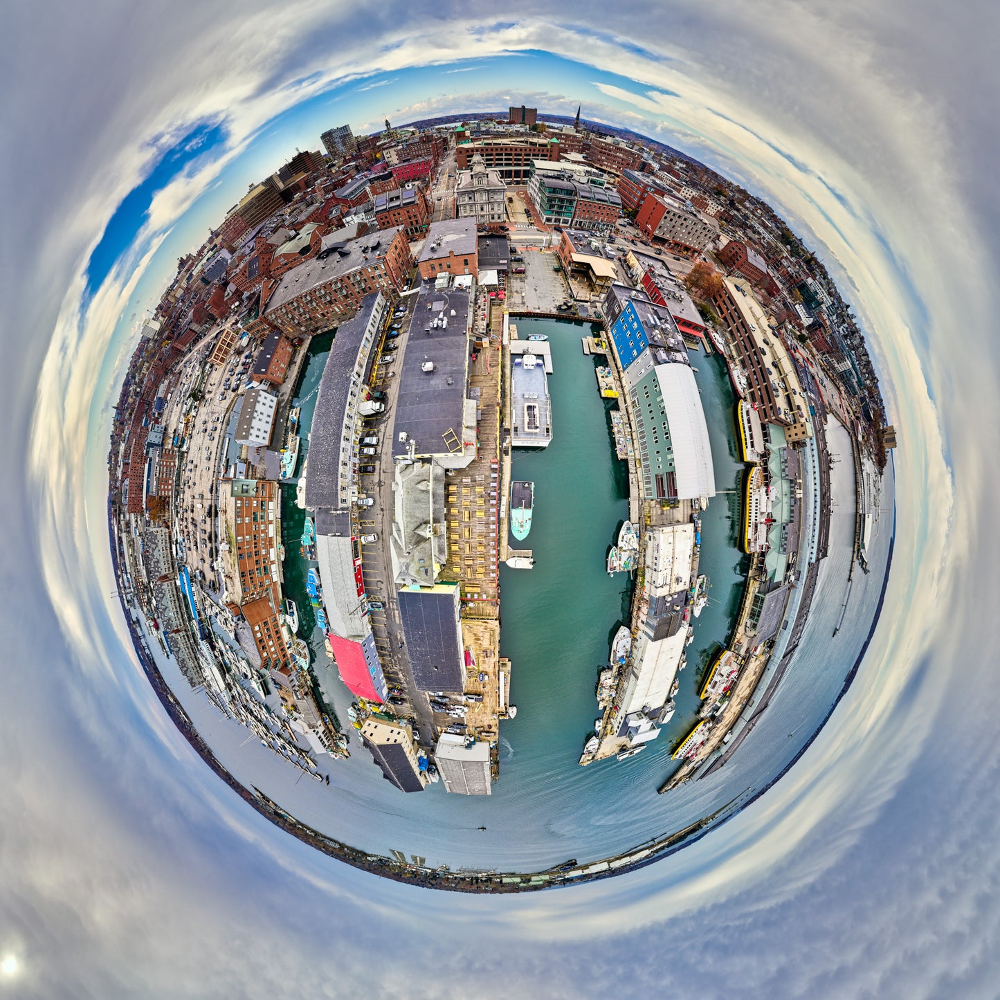 Alt text:
360-degree “tiny planet” image of Portland’s Old Port, with brick warehouses and long piers surrounding a central harbor basin filled with boats, all wrapped in a circular blue-gray sky.