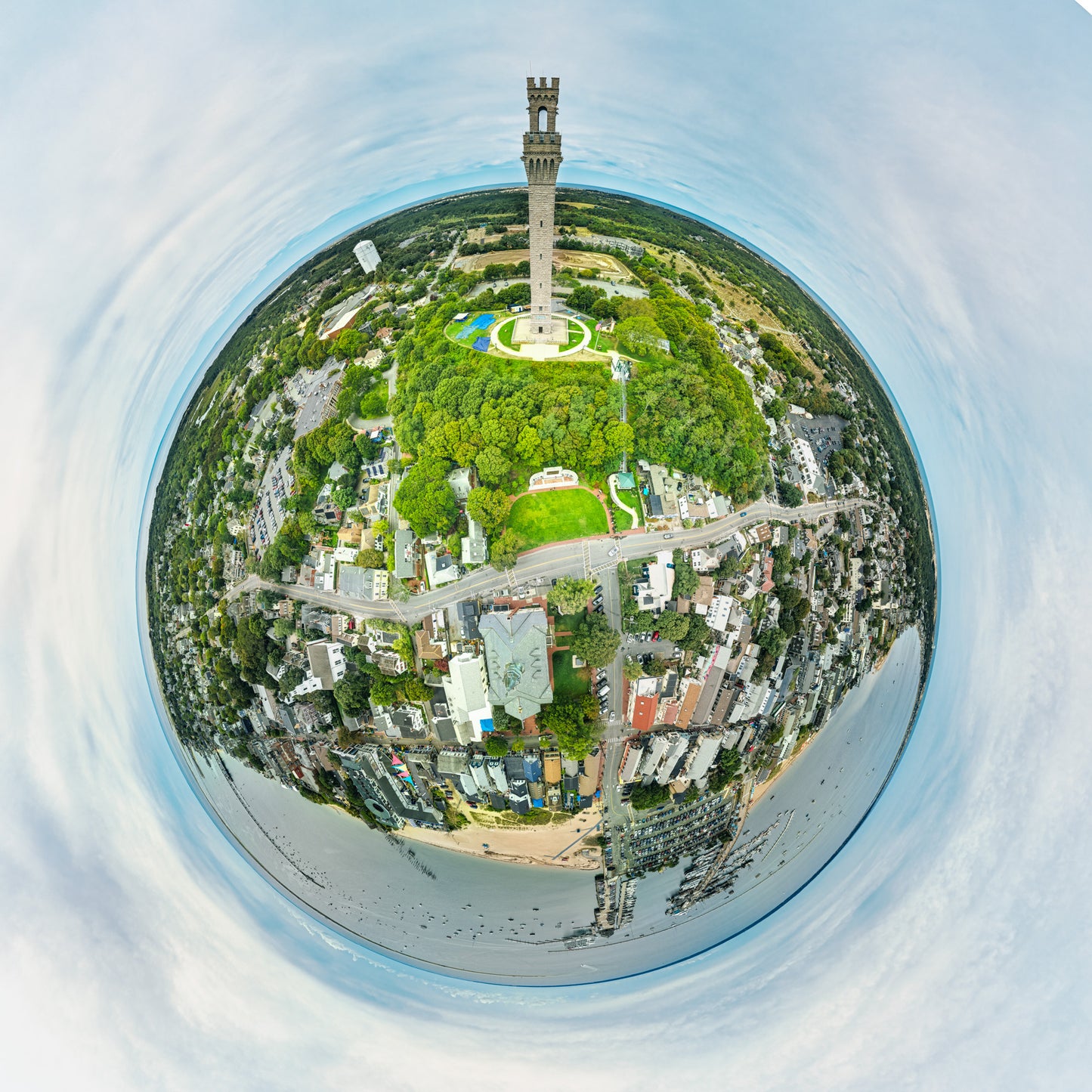 
360-degree “tiny planet” image of Provincetown, Massachusetts, with the tall Pilgrim Monument rising from a green hill at the center, surrounded by dense town streets, beaches, harbor with boats, and a circular pale blue sky.