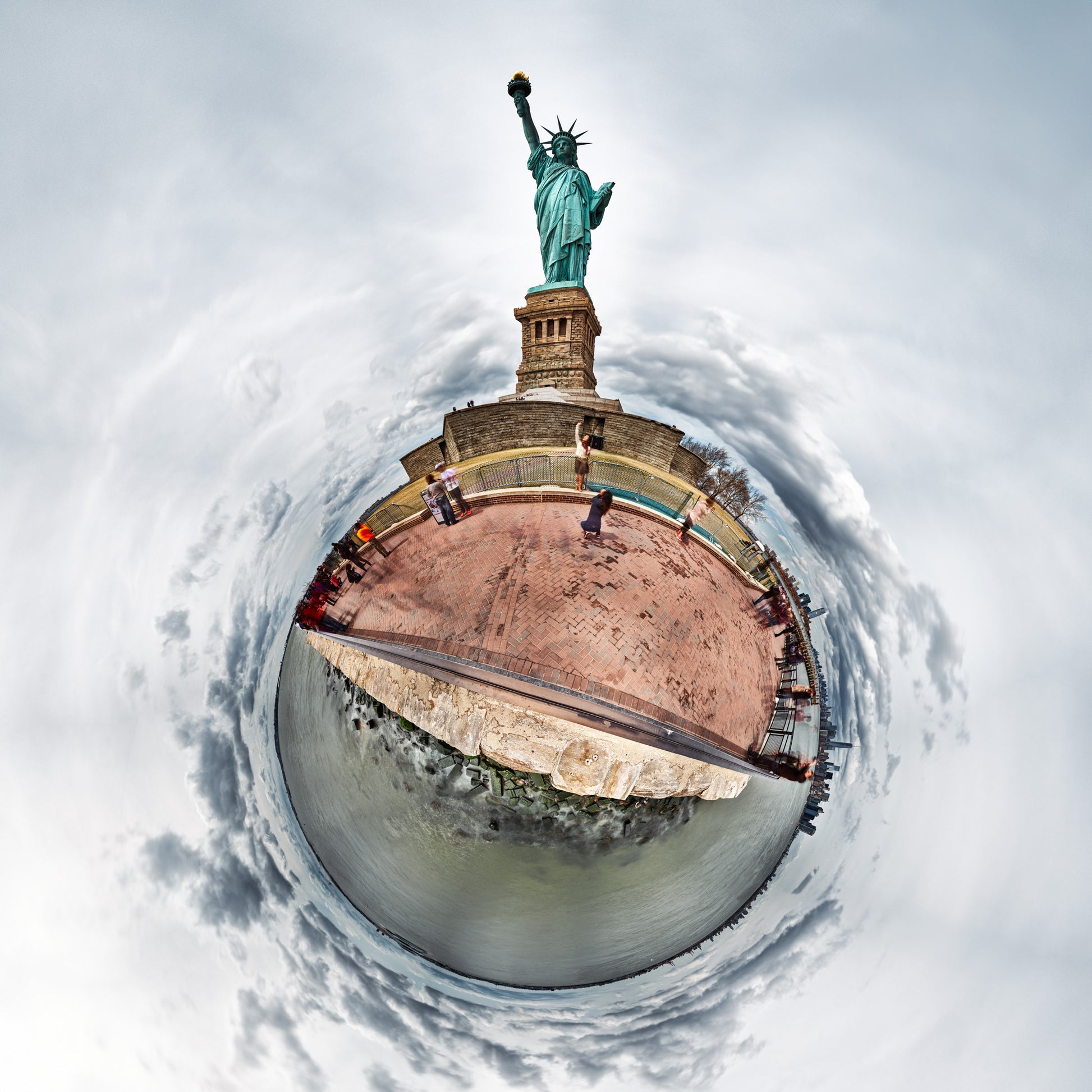 A 360-degree panoramic color image of the Statue of Liberty on an overcast afternoon, with a muted color palette of grays and greens, encircled by small figures against a continuous sky and water horizon